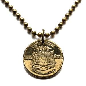 May include: A gold-toned circular coin pendant suspended from a matching ball chain necklace. The coin features an embossed emblem with intricate detailing, including rays at the top and Thai script characters surrounding the central design.