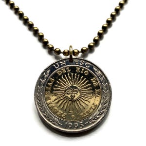Könnte beinhalten: Halskette mit einem Münzanhänger. Der Anhänger ist eine silber-goldene Münze mit dem Text "UN PESO" und "PROVINCIAS DEL RIO DE LA PLATA" um ein Sonnendesign. Die Kette hat eine bronzefarbene Kugelkette.