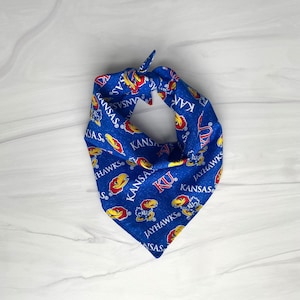 May include: Blue bandana with a repeating pattern of the word "KANSAS", the letters "KU", and a cartoon Jayhawk mascot in red and yellow. The bandana is tied at the top.