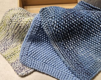 May include: Three knitted items in shades of blue, green, and white are displayed. The items appear to be hats or scarves, with a textured, handmade appearance. They are folded and arranged in a wooden box.
