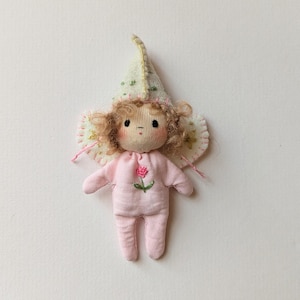 May include: A handmade doll with a pink jumpsuit and a flower embroidered on the front. The doll has curly brown hair, rosy cheeks, and a whimsical hat with green and gold accents. The doll's wings are a light cream color with pink stitching.