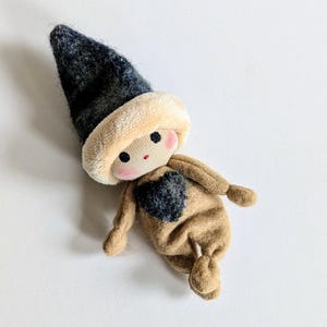 May include: A small, handmade doll with a brown felt body, a black felt heart on its chest, and a black and white felt hat. The doll has a stitched face with red cheeks and black eyes.