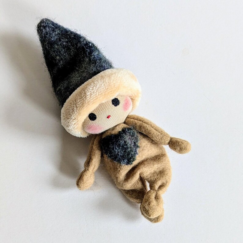 May include: A small, handmade doll with a brown felt body, a black felt heart on its chest, and a black and white felt hat. The doll has a stitched face with red cheeks and black eyes.