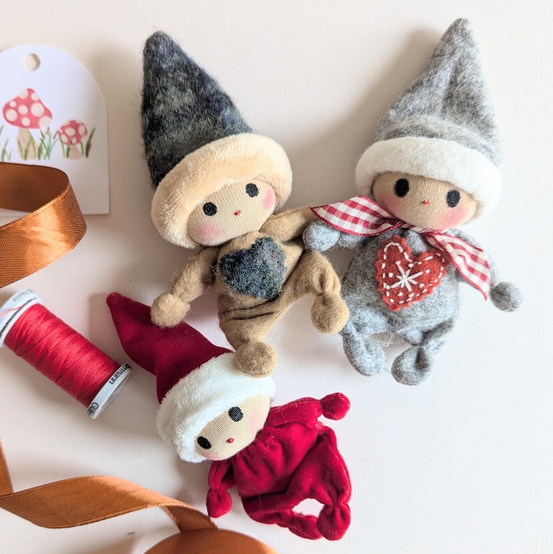 May include: Three handmade felt Christmas elf dolls. One elf is wearing a gray hat and a gray outfit with a red heart on the front. Another elf is wearing a brown hat and a brown outfit with a gray heart on the front. The third elf is wearing a red hat and a red outfit.
