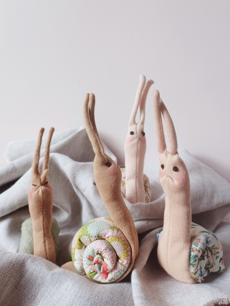 May include: Four handmade fabric snails in various shades of brown and pink. The snails have floral patterned shells and are arranged on a light gray fabric background.