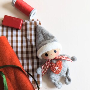 May include: A gray felt elf doll with a white felt hat and a red heart on its chest. The doll is sitting on a brown and white checkered fabric.