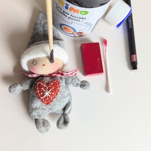 May include: A gray felt doll with a red heart on its chest and a white hat. The doll is being painted with a black paint brush. The image also shows a bottle of black fabric paint, a red wooden block, and a cotton swab.