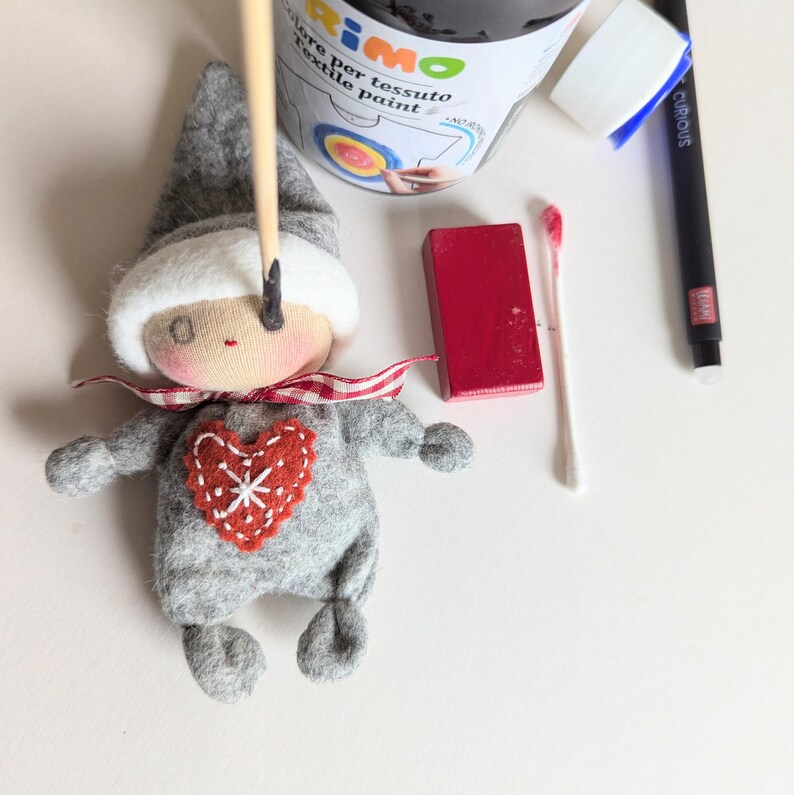 May include: A gray felt doll with a red heart on its chest and a white hat. The doll is being painted with a black paint brush. The image also shows a bottle of black fabric paint, a red wooden block, and a cotton swab.