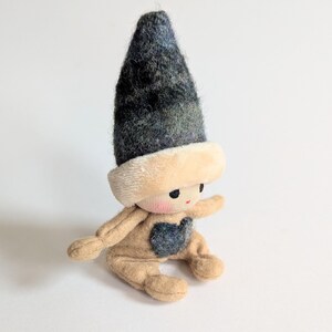 May include: A small, handmade doll with a tan felt body and a black and blue felt hat. The doll has a heart-shaped patch on its chest and a stitched face with black eyes and a red mouth.