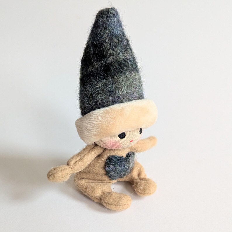May include: A small, handmade doll with a tan felt body and a black and blue felt hat. The doll has a heart-shaped patch on its chest and a stitched face with black eyes and a red mouth.