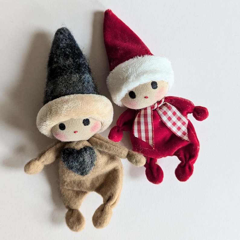 May include: Two handmade felt Christmas elf dolls. One elf is wearing a brown felt outfit with a black heart and a black and white hat. The other elf is wearing a red felt outfit with a white and red gingham ribbon and a red and white hat.
