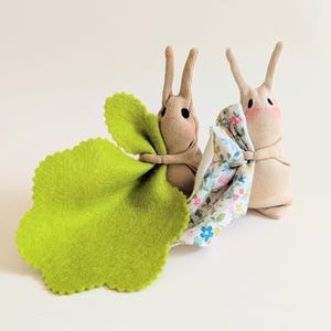 Peut inclure: Deux poupées lapins faites à la main, aux joues roses et aux longues oreilles. L'un des lapins tient une grande feuille de feutre vert vif, tandis que l'autre tient un tissu à motifs floraux. Les lapins sont en tissu beige.