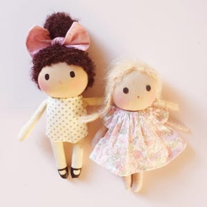 May include: Two handmade cloth dolls with embroidered faces. One doll has brown hair with a pink bow and a white dress with brown polka dots. The other doll has blonde hair and a white dress with a floral pattern.