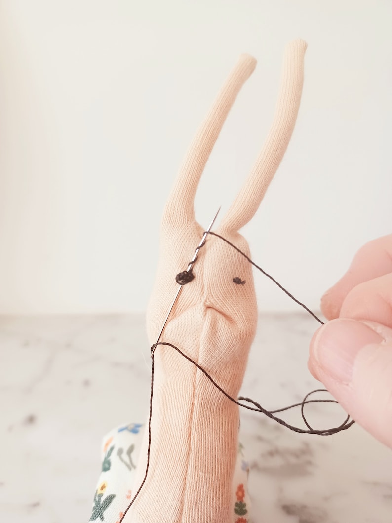 May include: A close-up of a light pink fabric snail with black eyes being sewn with a needle and black thread. The snail is sitting on a fabric with a floral pattern.