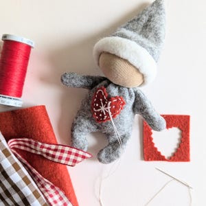 May include: A gray felt doll with a white felt hat and a red felt heart on its chest. The doll is sitting on a white surface with red felt scraps and a needle and thread.