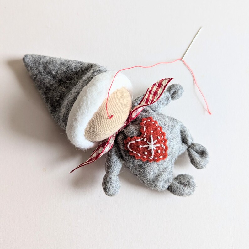 May include: A gray felt gnome with a white felt hat, a red and white gingham ribbon, and a red felt heart with a white snowflake embroidered on it. The gnome is being sewn together with red thread.