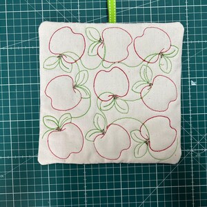 May include: A square, off-white fabric item with a stitched apple design in red and green. The apples are outlined with red thread and have green leaves. A green ribbon is attached at the top. The item is on a green cutting mat.