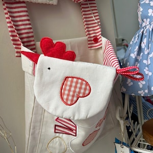 May include: A hanging fabric storage organizer featuring a white chicken-shaped pocket with a red gingham heart applique. The organizer has red and white striped straps and a top panel decorated with embroidered apple shapes and red stitching.