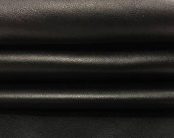 May include: A piece of black leather fabric featuring a distinct, fine-grained texture. The material is folded into several horizontal layers, creating deep shadows and highlights that emphasize the matte finish and the supple quality of the surface.