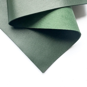 Op de afbeelding: Close-up van twee vellen groen leer. Het bovenste vel is omgevouwen en onthult een lichtere, suède-achtige onderkant. De bovenste laag heeft een getextureerd, korrelig oppervlak. Het onderste vel is een donkerdere tint groen.