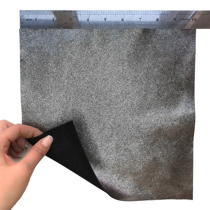 May include: A piece of silver-colored faux leather with a textured surface, approximately 9 inches long. The back of the material is black. A metal ruler is placed across the top of the material.