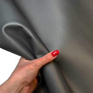 May include: A close-up of a sheet of dark gray leather. The leather is smooth and appears to be folded. The image is well-lit, highlighting the texture and color of the material. The leather is being held by a hand with red nail polish.