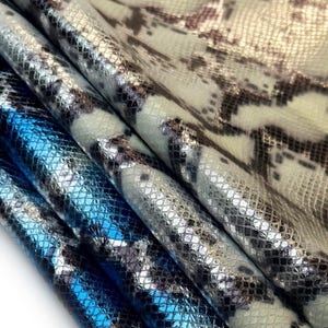 Può includere: Primo piano di tre tessuti strutturati con un motivo a pelle di serpente. I tessuti sono in tonalità di blu, argento e verde chiaro con accenti marrone scuro. I tessuti sono piegati l'uno sull'altro, creando un effetto a strati.