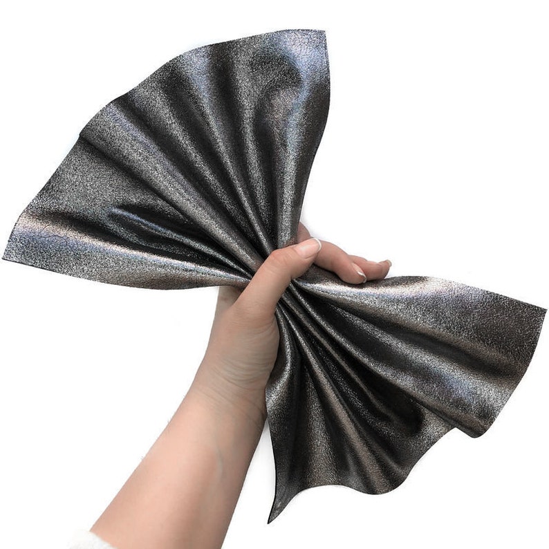 May include: A piece of metallic silver fabric, held in a hand, fanned out to display its texture and sheen. The fabric has a textured surface and a dark silver color, with hints of other colors.