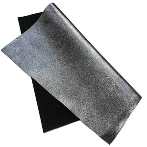 May include: A sheet of silver glitter fabric layered over a black sheet. The silver fabric has a textured, speckled appearance, and is slightly curled at the top. The black fabric is smooth and matte.