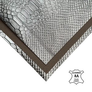Puede incluir: Primer plano de hojas de cuero plateado con un patrón de escamas de reptil. El cuero está superpuesto a una hoja marrón. El logotipo de Italian Leather Company está en la esquina inferior derecha.