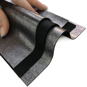 May include: Close-up of layered leather samples. The top layer is a metallic silver with a textured finish, revealing a black layer beneath. The bottom layer is a solid black leather. The samples are being held up, showcasing the different textures.
