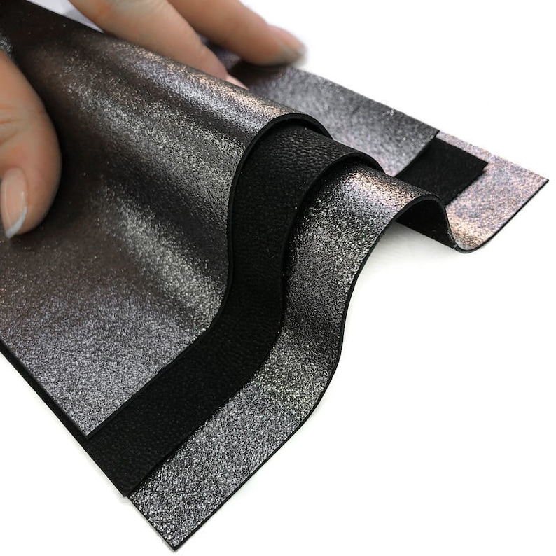 May include: Close-up of layered leather samples. The top layer is a metallic silver with a textured finish, revealing a black layer beneath. The bottom layer is a solid black leather. The samples are being held up, showcasing the different textures.