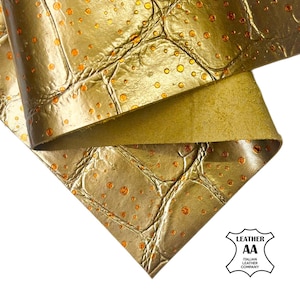 May include: Close-up of gold-colored leather with a crocodile skin texture. The leather has a metallic sheen and is adorned with small, orange-colored circular accents. The underside of the leather is a solid, lighter gold color. The image includes a logo.