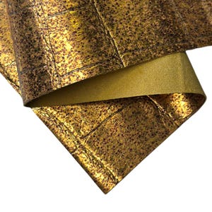May include: Close-up of a folded piece of gold-colored leather with a textured, mottled surface. The leather has a metallic sheen and a grid-like pattern, with a contrasting lighter gold underside visible.