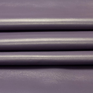 Pode incluir: Close-up de uma folha de couro dobrada na cor lavanda. O couro tem uma superfície lisa e ligeiramente brilhante com dobras e vincos visíveis. A cor é um roxo suave, com variações sutis de tom.