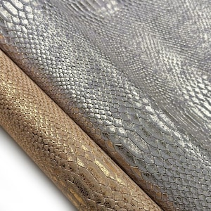 May include: Close-up of two rolls of textured faux leather. One roll is silver, and the other is gold. The surface of each roll has a scale-like pattern. The rolls are angled, showing the texture and sheen of the material.