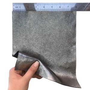 May include: A piece of silver glitter fabric is held up to a ruler. The fabric is approximately 8 inches wide. The fabric has a textured surface and a shiny appearance. The ruler is marked in inches.