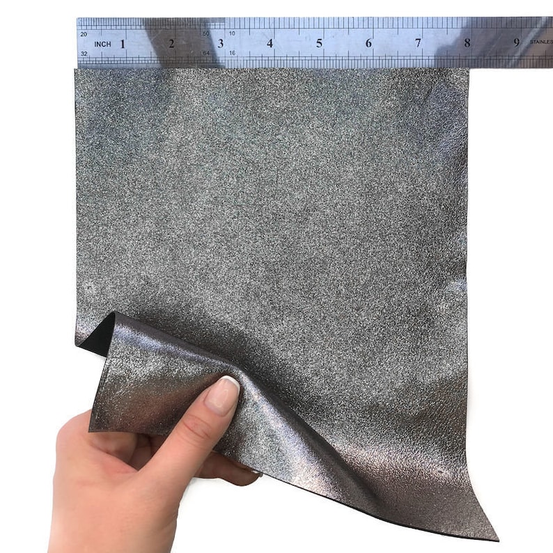 May include: A piece of silver glitter fabric is held up to a ruler. The fabric is approximately 8 inches wide. The fabric has a textured surface and a shiny appearance. The ruler is marked in inches.