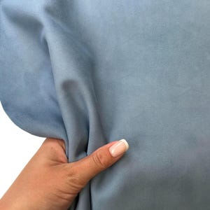 Op de afbeelding: Close-up van een lichtblauwe stof met een zachte, fluweelachtige textuur. De stof wordt vastgehouden en toont de drape en subtiele kleurvariaties. Het materiaal lijkt glad en mogelijk geschikt voor kleding of woondecoratie.