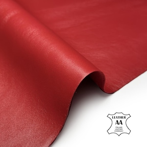 Puede incluir: Una pieza de cuero rojo vibrante doblada para mostrar su textura suave de grano fino y acabado mate. Un logotipo blanco en la esquina inferior derecha presenta el texto LEATHER AA ITALIAN LEATHER COMPANY dentro de un contorno en forma de escudo.