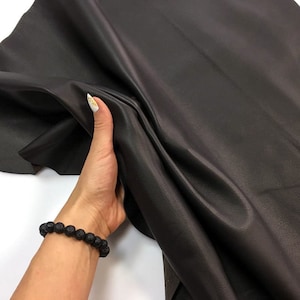 May include: A close-up of a dark brown leather hide. The leather is being held up by a hand, showing its texture and flexibility. The leather appears to be smooth and has a slight sheen.