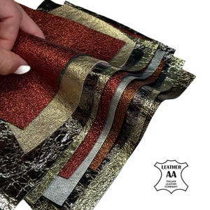 Puede incluir: Una pila de láminas de cuero superpuestas con acabados metálicos en rojo, oro, plata y negro. Cada pieza presenta una superficie texturizada y arrugada. Un logotipo en la esquina inferior derecha dice LEATHER AA ITALIAN LEATHER COMPANY.