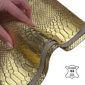 Könnte beinhalten: Nahaufnahme von goldfarbenem Leder mit Schlangenhautmuster. Das Leder wird angehoben, um die Unterseite und die Schichten freizulegen. Das Bild enthält ein Logo für ein italienisches Lederunternehmen.