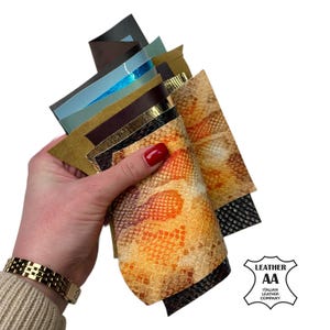 Puede incluir: Una mano sostiene una colección de muestras de cuero en varios colores y texturas. Las muestras incluyen tonos de marrón, dorado, negro y azul, con algunos diseños metálicos y de piel de serpiente. El logotipo de Italian Leather Company es visible.