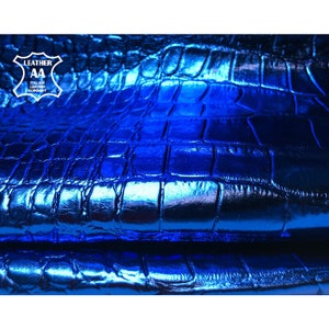 Könnte beinhalten: Ein Stück metallisch blaues Leder mit einer erhabenen, geprägten Krokodilleder-Struktur. Die Oberfläche weist ein glänzendes, reflektierendes Finish in tiefen Blautönen auf. Ein weißes Logo oben links zeigt LEATHER AA ITALIAN LEATHER COMPANY.