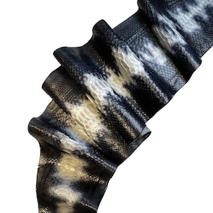 May include: Close-up of a folded piece of snake skin. The skin has a pattern of black, white, and gray scales. The scales are small and tightly packed together, creating a textured surface. The skin is folded over itself, revealing the underside.