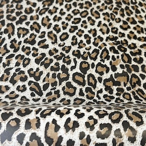 Pode incluir: Close-up de um tecido com um padrão de estampa de leopardo. O padrão apresenta manchas pretas, marrons e bege em um fundo creme. O tecido parece estar dobrado, mostrando a textura e a profundidade do material.