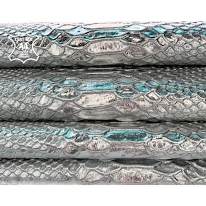 Peut inclure: Gros plan sur des feuilles de cuir empilées et texturées avec un motif métallique en forme de peau de serpent. La couleur est principalement argentée avec des touches de sarcelle et une finition brillante. Le cuir a un motif en forme d'écailles.