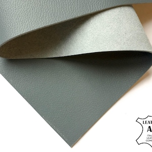 Puede incluir: Primer plano de una lámina de cuero gris con superficie texturizada, parcialmente enrollada para revelar un reverso gris más claro. El logotipo de Italian Leather Company está en la esquina inferior derecha.