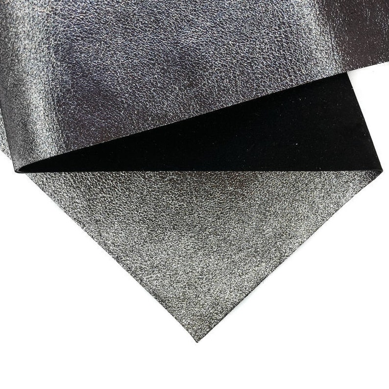 May include: Close-up of a folded piece of silver metallic fabric with a textured surface. The underside of the fabric is black. The fabric is on a white background.
